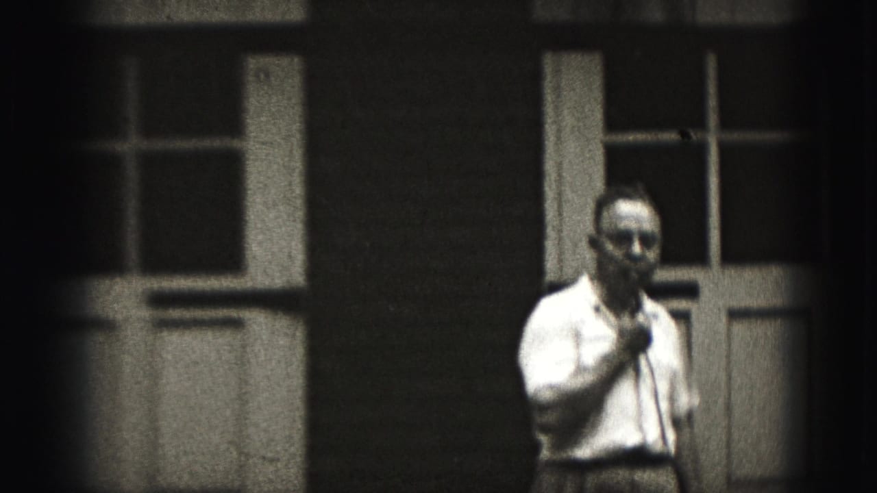 Man in a light shirt holds a microphone outside paneled doors.