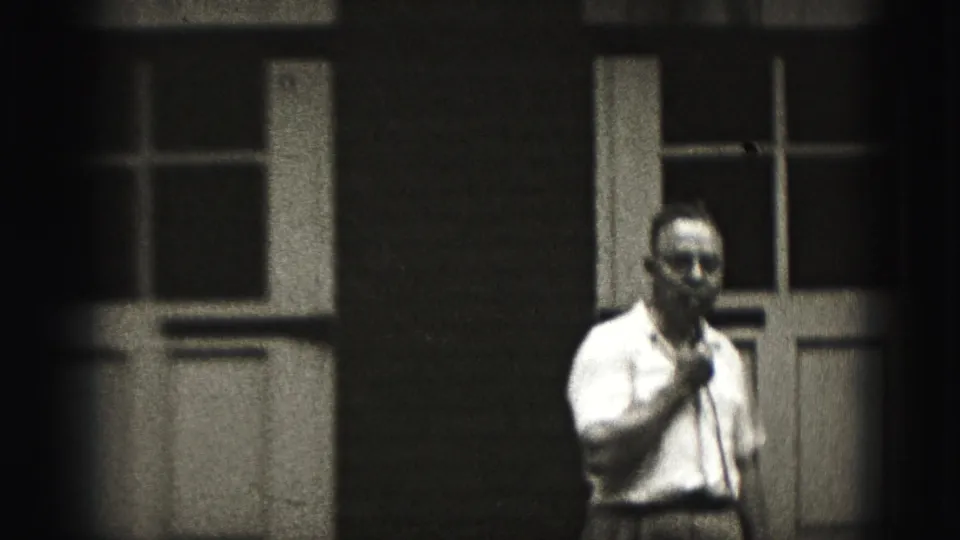 In 1975 Harrisburg footage, a man in a light short-sleeved shirt stands outside a building holding a handheld microphone.