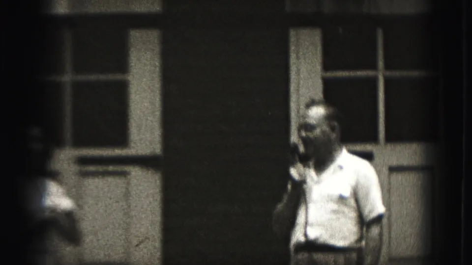 The performer is seen in partial profile with a handheld microphone near paneled doors in the Harrisburg home-movie footage.