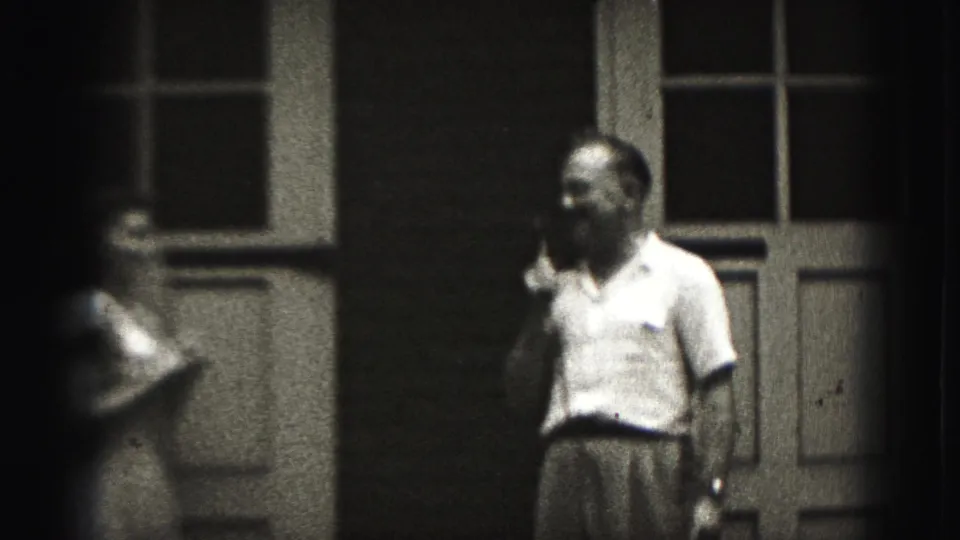 In 1975 Harrisburg home-movie footage, a man with a microphone stands by paneled doors as a girl moves near the left edge.