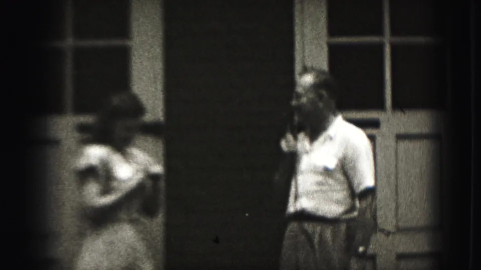 A girl and a man with a handheld microphone stand outside a Harrisburg house in grainy 1975 8mm footage.