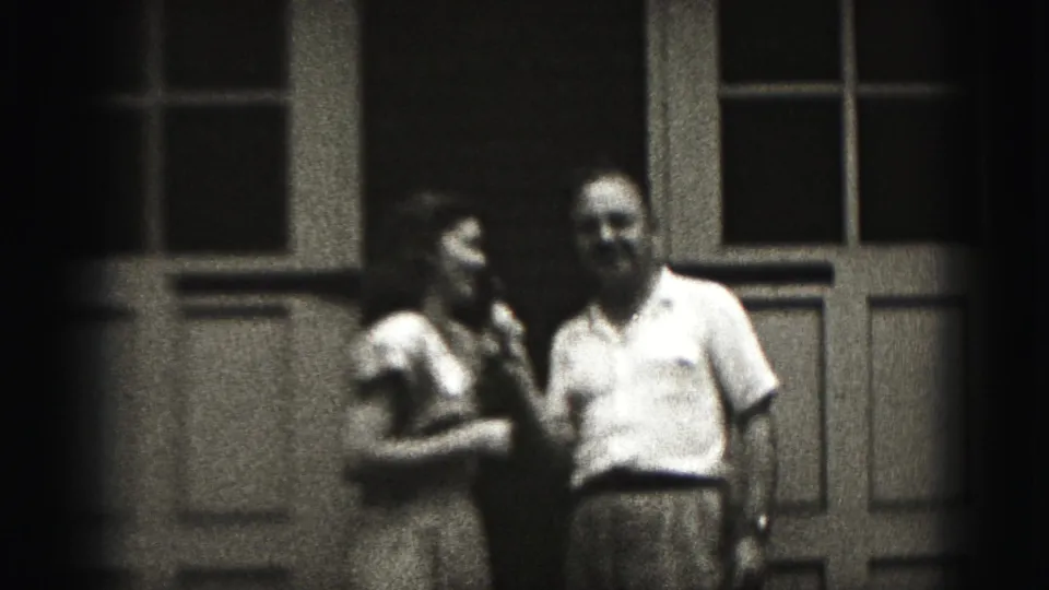 In 1975 Harrisburg home-movie footage, a woman and man stand before paneled doors as she holds a microphone near them.