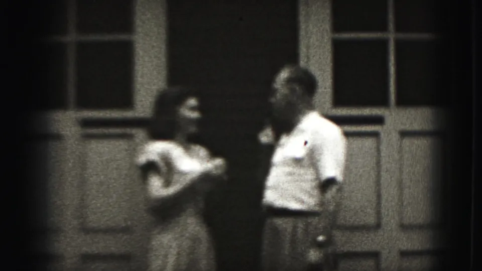 The woman turns toward the man during the microphone exchange, with paneled doors and window panes behind them in the 1975 home movie.