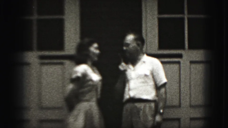 A woman in a light dress and a man in a light shirt converse outside a paneled doorway in 1975 Harrisburg footage.