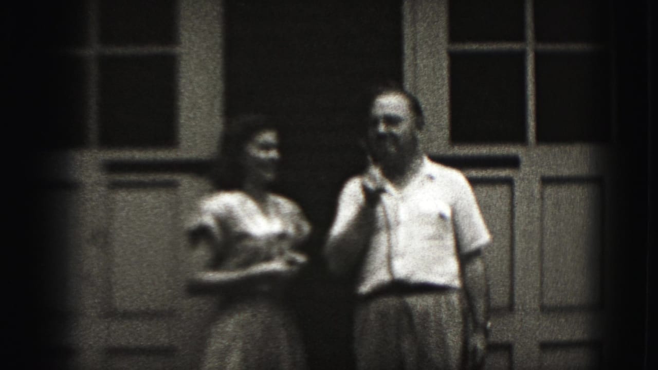 In 1975 Harrisburg home-movie footage, a man holds a microphone beside a woman on a shaded porch with tall windows and paneled doors.