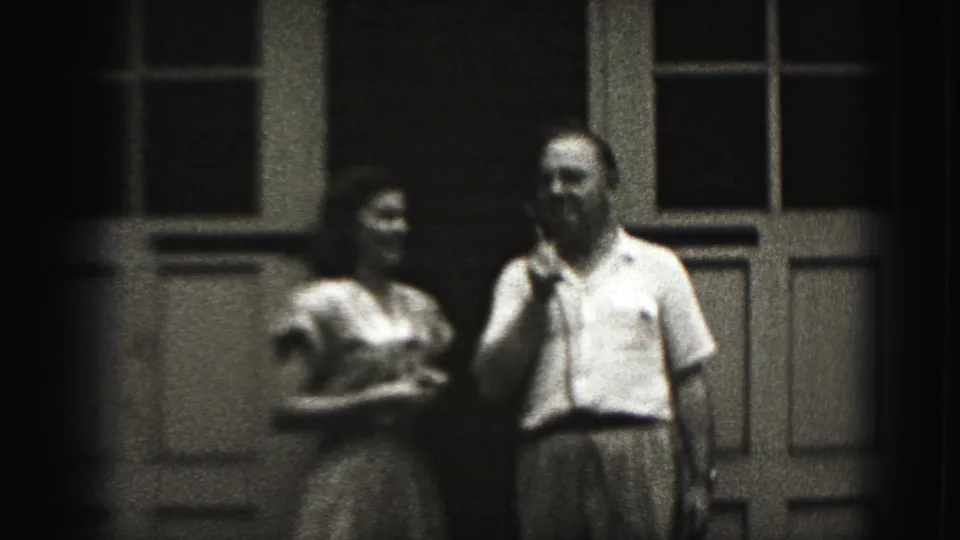 In 1975 Harrisburg home-movie footage, a man holds a microphone beside a woman on a shaded porch with tall windows and paneled doors.