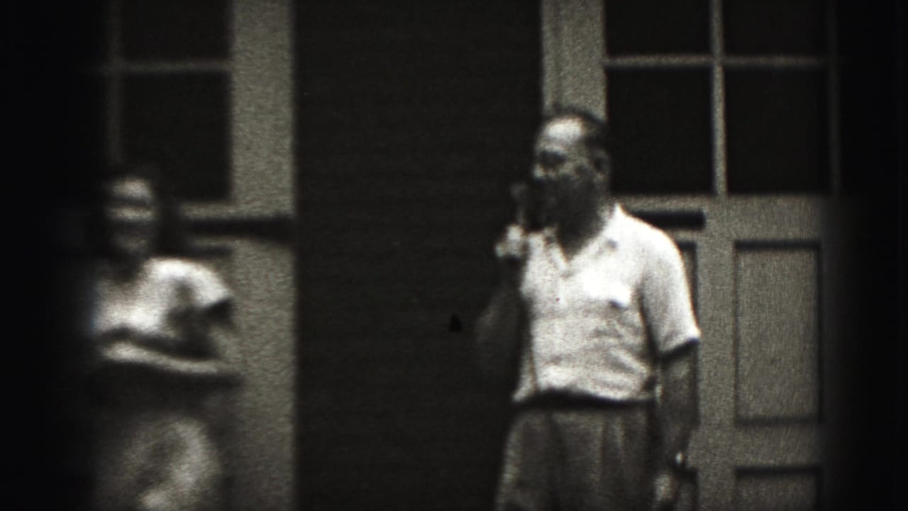 A man in a light shirt holds a microphone while a blurred woman stands nearby in the 1975 Harrisburg porch scene.