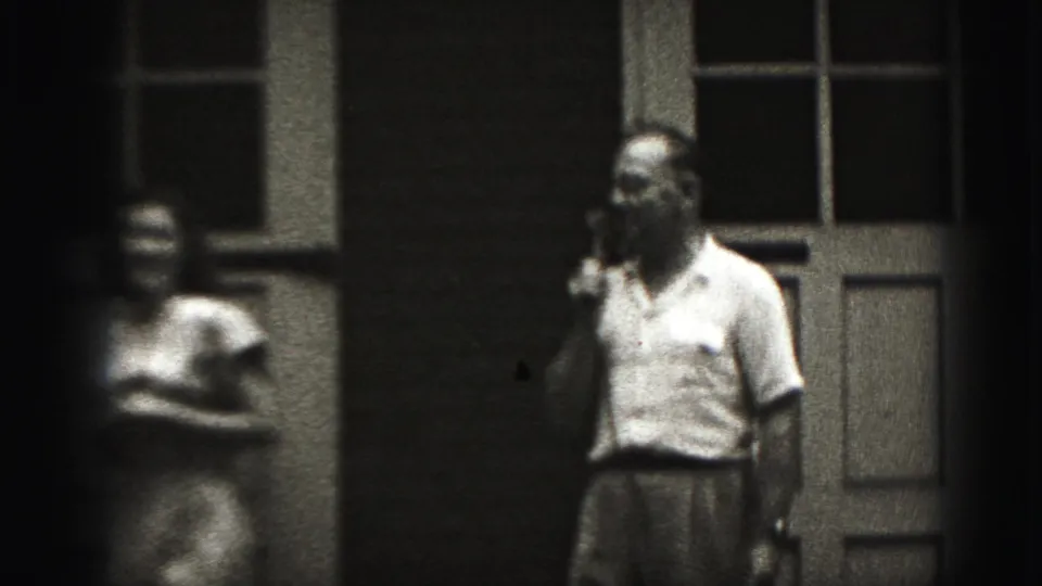 A man in a light shirt holds a microphone while a blurred woman stands nearby in the 1975 Harrisburg porch scene.