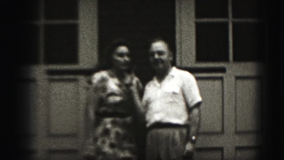 In metadata-supported 1975 Harrisburg footage, a woman in a patterned dress and a man in a light shirt stand before paneled doors.
