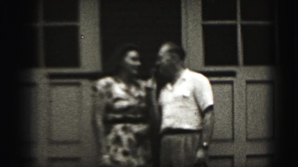 A man in a light shirt leans toward a woman in a patterned dress during a brief exchange in the 1975 Harrisburg home-movie scene.