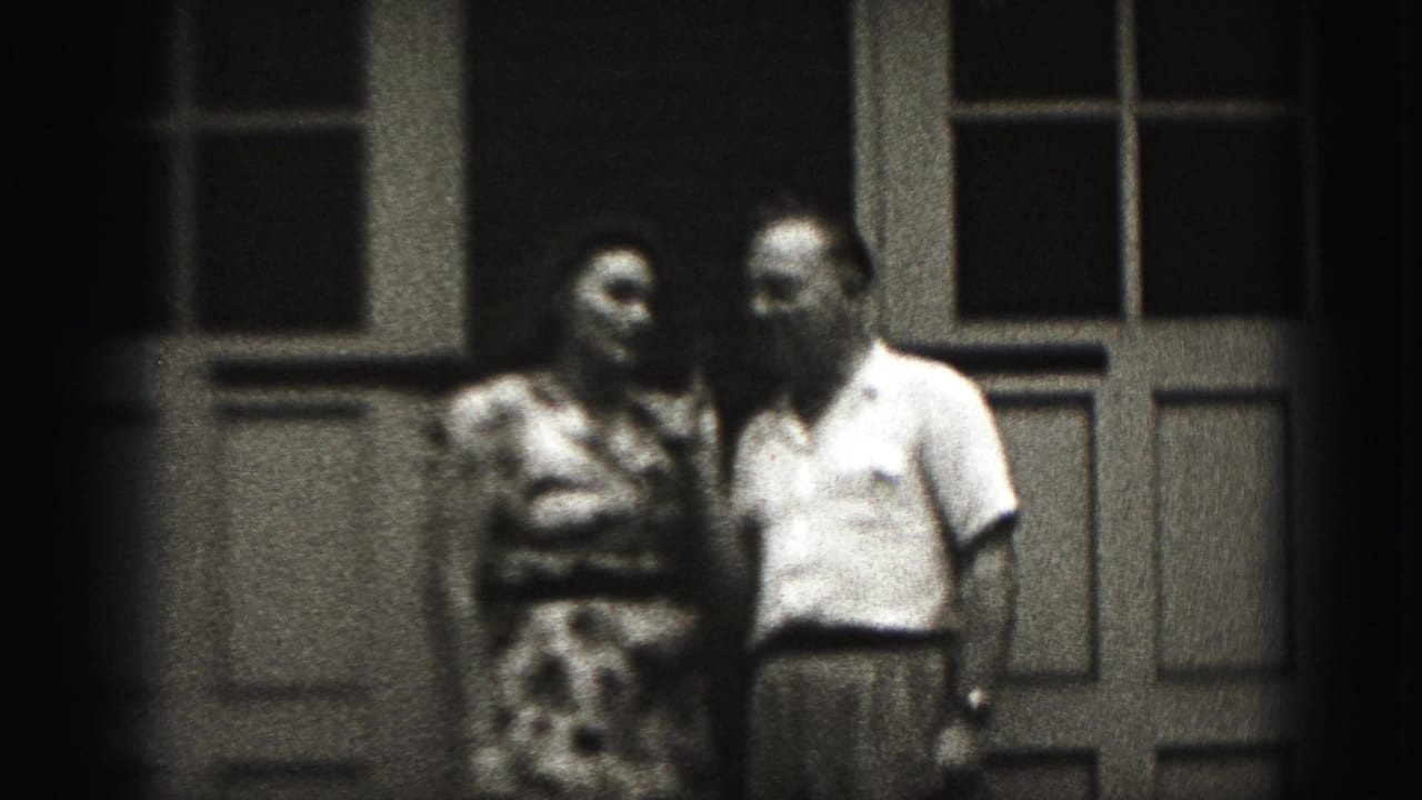 In Harrisburg, Pennsylvania home-movie footage from 1975, an adult couple stands close on a porch before a paneled door and windows.