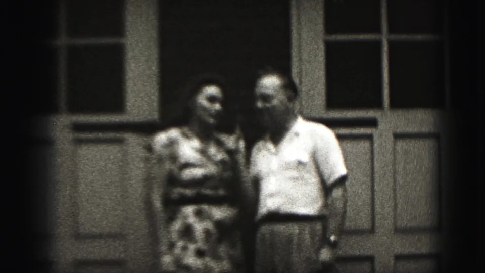 In Harrisburg, Pennsylvania home-movie footage from 1975, an adult couple stands close on a porch before a paneled door and windows.