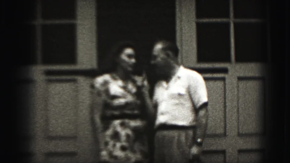 In 1975 Harrisburg home-movie footage, a man with a microphone stands beside a woman in a patterned dress outside paneled doors.