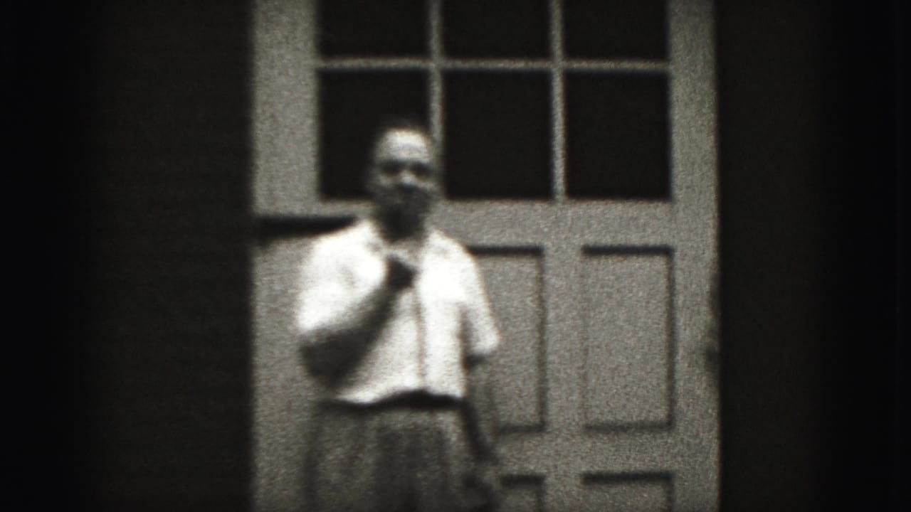 In 1975 Harrisburg home-movie footage, an adult man in a white shirt stands before a paneled door, hand raised near his chin.