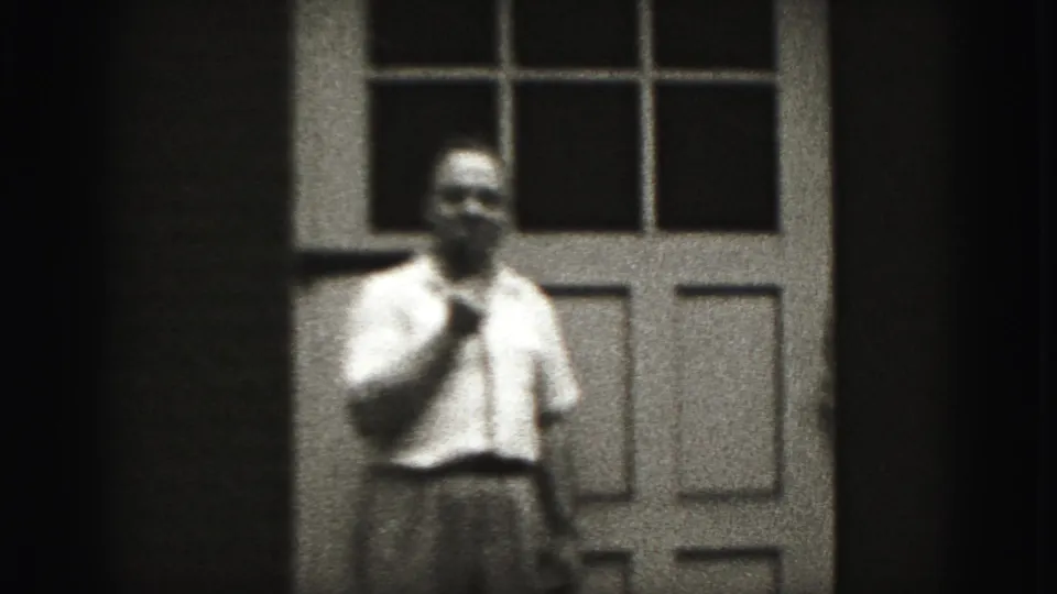 In 1975 Harrisburg home-movie footage, an adult man in a white shirt stands before a paneled door, hand raised near his chin.