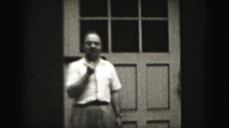 The man remains near the doorway in Harrisburg, facing forward with a contemplative hand gesture in grainy 8mm footage.