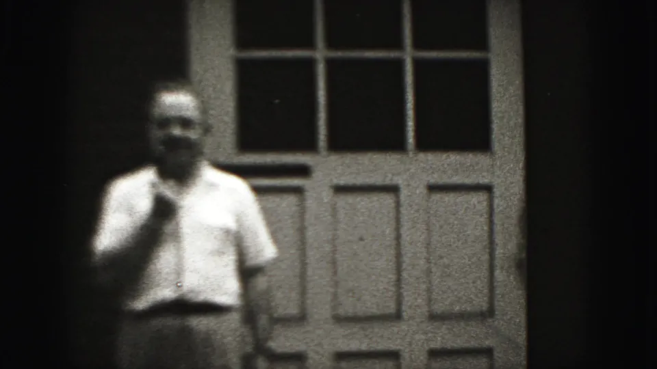 The 1975 home-movie frame shows the man standing in front of the paneled door, his face and upper body softly rendered by the 8mm scan.
