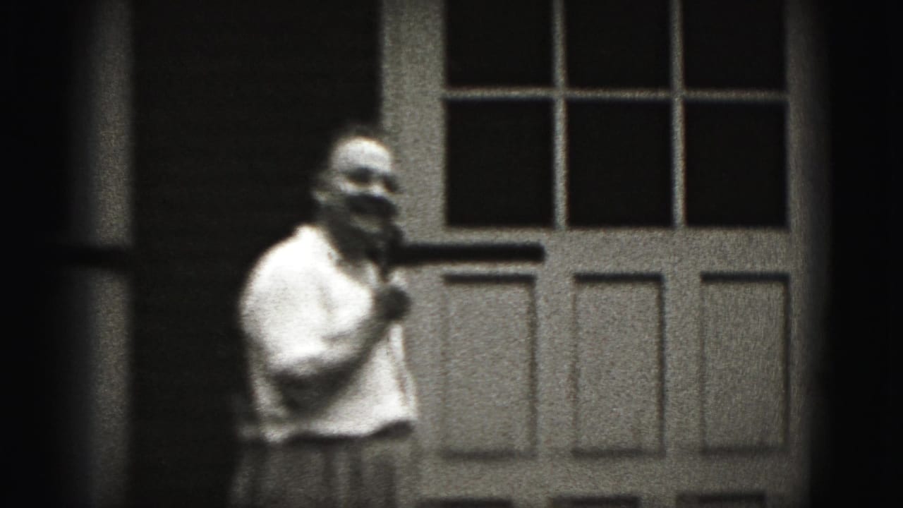 Man in a white shirt holding a microphone beside a paneled door.