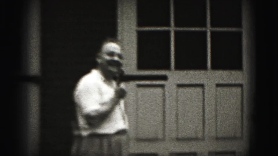 Adult man in a white shirt speaks into a microphone near a paneled door in 1975 Harrisburg home-movie footage.