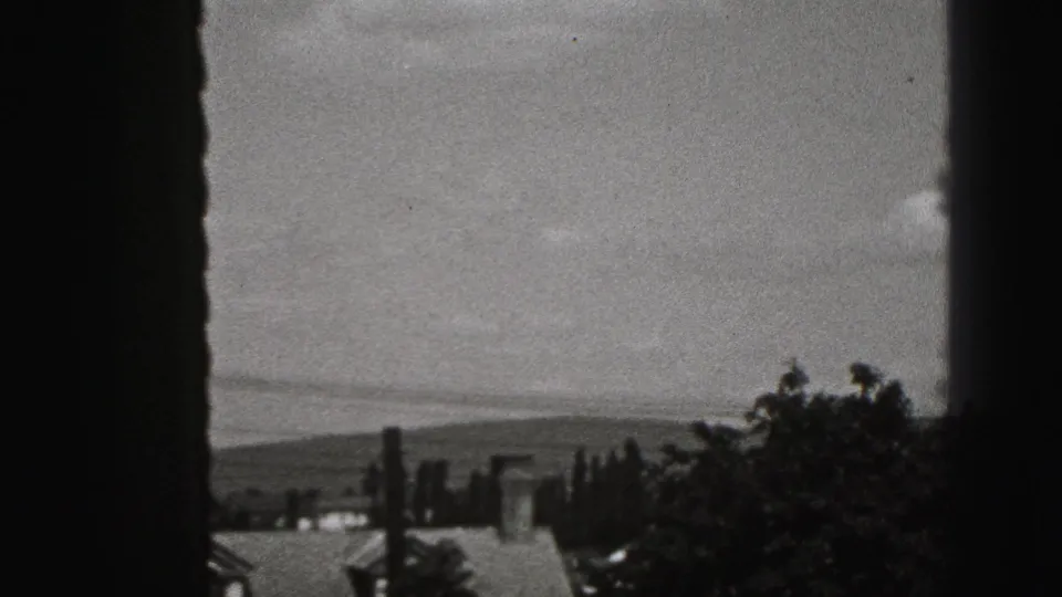 A dark window frame divides a soft 8mm view of rooftops, trees, and distant hills in Harrisburg.