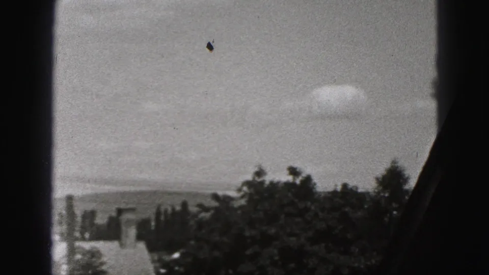 The final frame holds on rooftops, trees, cloudy sky, and distant hills through a dark window edge.