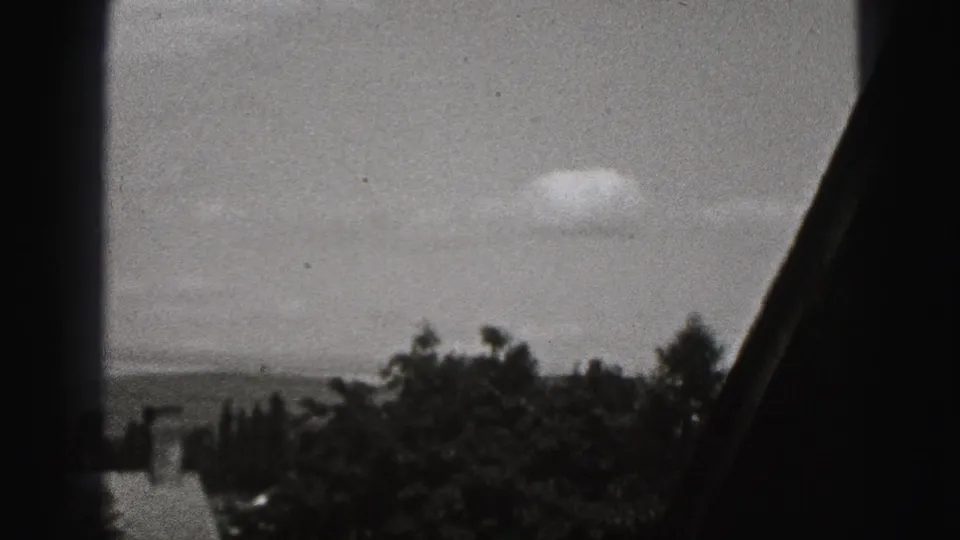1975 Harrisburg home-movie frame looking through a window toward cloudy sky and trees beyond a residential exterior.