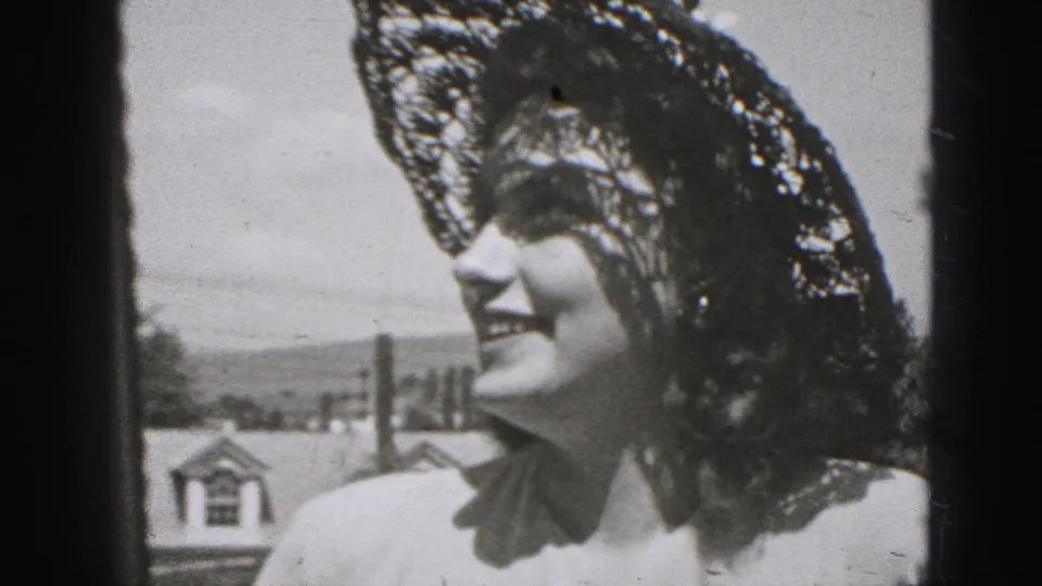 Young woman in a delicate lace hat smiles outdoors in 1975 Harrisburg home-movie footage, with rooftops and sky behind her.