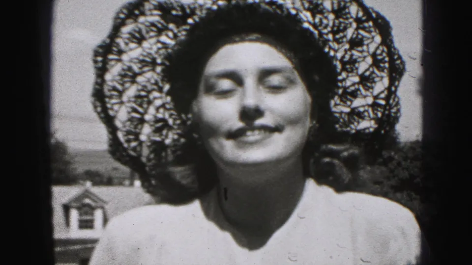Woman faces the camera with eyes closed under a lace-style sun hat in 1975 Harrisburg home-movie footage.
