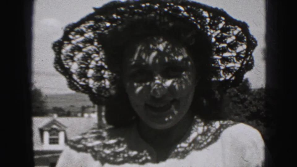 Woman under a lace-style hat faces the camera in shadow, with outdoor buildings and trees behind her in Harrisburg, 1975.