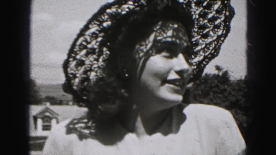 The woman smiles gently beneath a wide lace hat in 1975 Harrisburg home-movie footage, with trees and a roofline behind her.