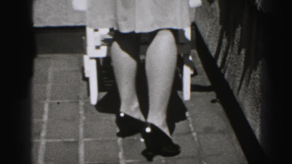 In 1975 Harrisburg home-movie footage, a woman in a light dress and dark shoes walks past a white outdoor chair in sunlight.