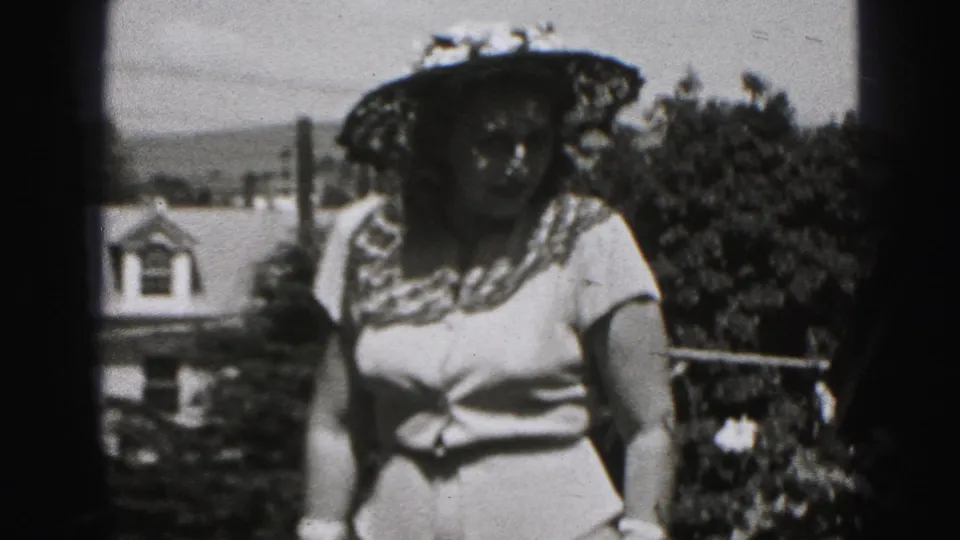 A woman in a floral wide-brim hat and light dress faces the home-movie camera in a sunny Harrisburg outdoor setting.