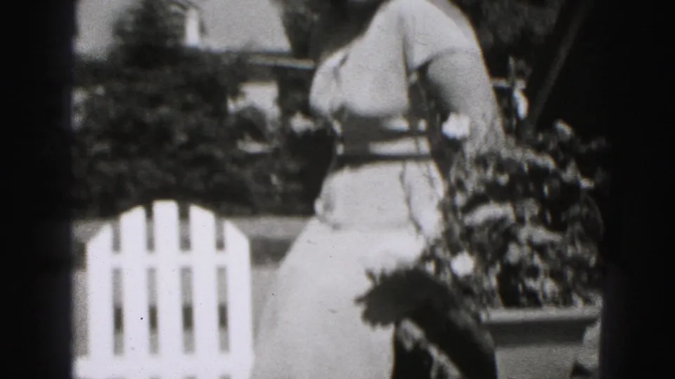 The woman in a light dress is framed beside a tree trunk and white fence in 1975 Harrisburg home-movie footage.