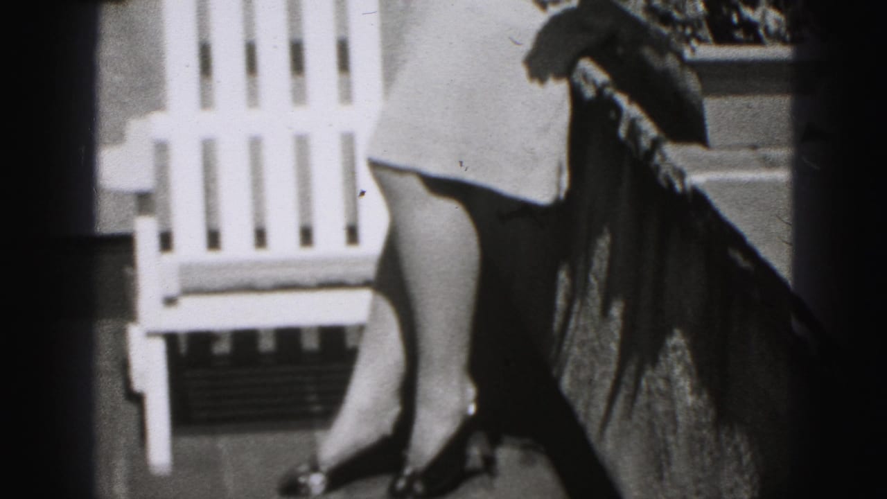 1975 Harrisburg home-movie frame showing a porch railing, white Adirondack chair and part of a woman in a wide hat.