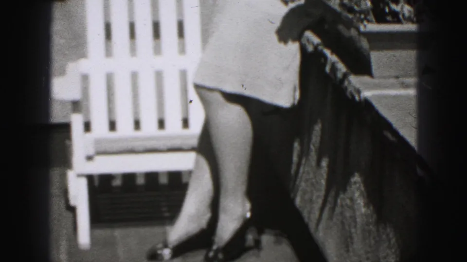 1975 Harrisburg home-movie frame showing a porch railing, white Adirondack chair and part of a woman in a wide hat.