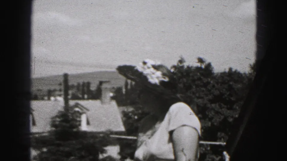 A woman in a wide-brimmed hat stands on a porch in Harrisburg, Pennsylvania, with neighborhood rooftops behind her.