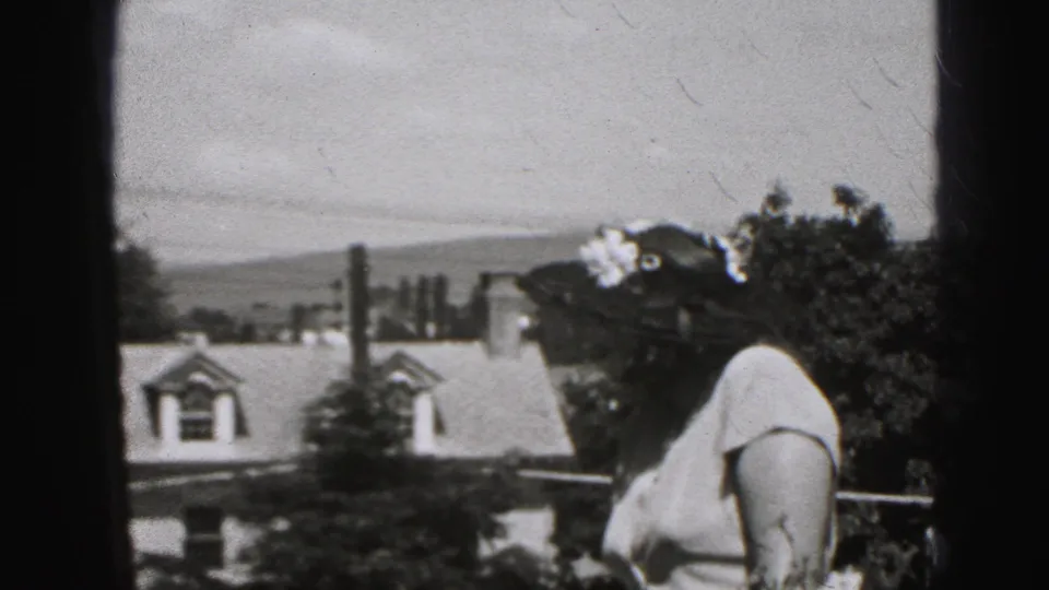 Side view of the woman in a broad sunhat and light dress on a Harrisburg porch, with houses and trees beyond.
