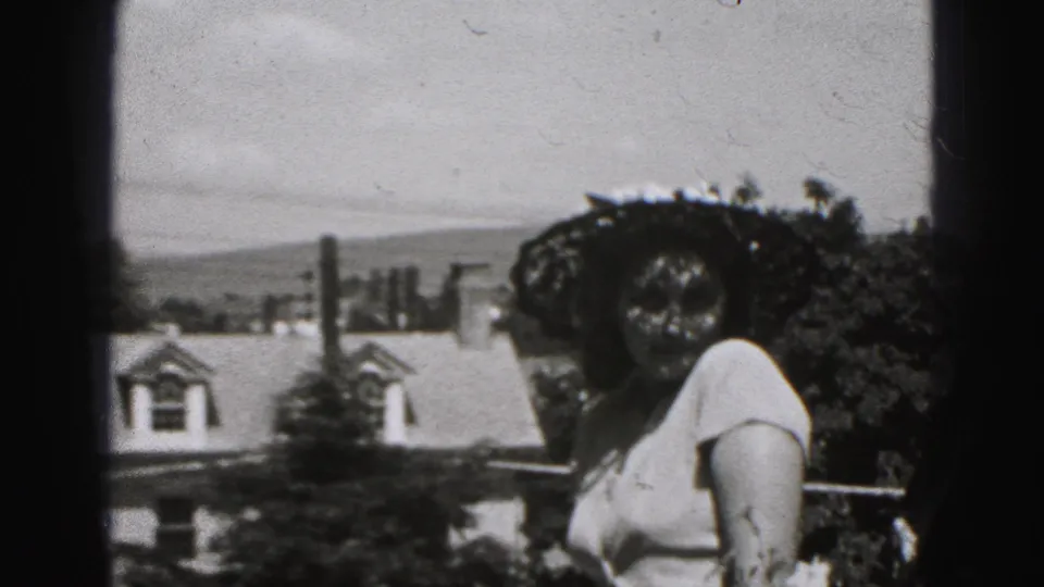 1975 Harrisburg home-movie frame showing a woman in a large dark hat making a face near residential rooftops and trees.