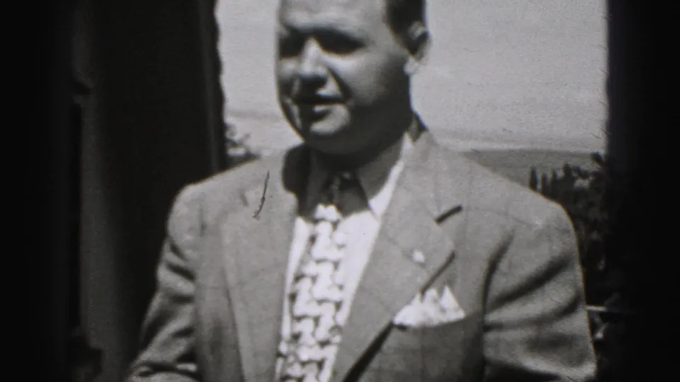 1975 Harrisburg home-movie frame showing a suited man with patterned tie and pocket square outdoors near residential rooftops.