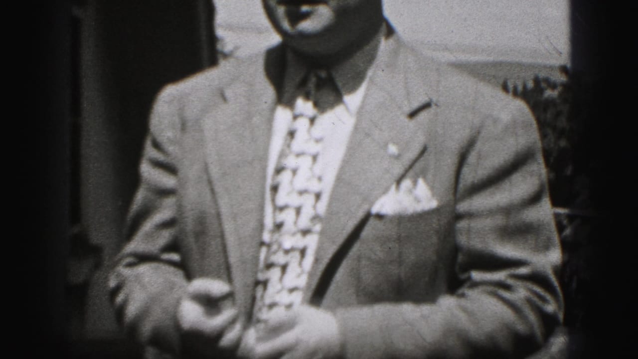 Adult man in a suit and patterned tie stands partly cropped beside a dark doorway.