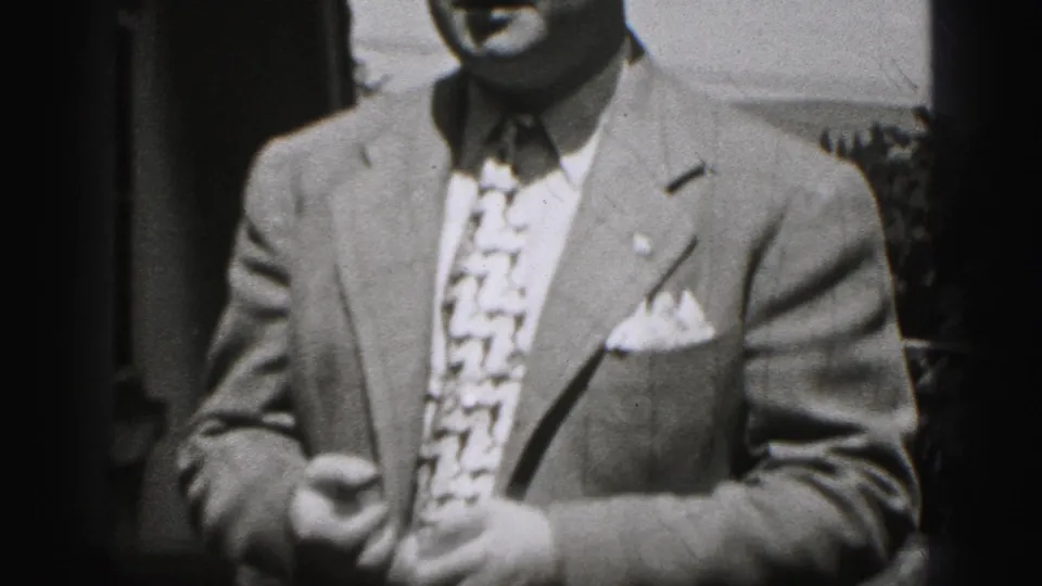 In this 1975 Harrisburg home-movie frame, a suited adult man with a patterned tie is partly cropped near a residential doorway.