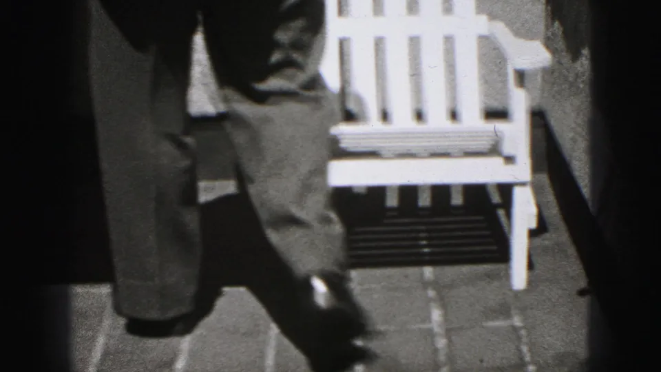 A low, cropped view shows the suited man stepping past a white chair on patio paving in the 1975 Harrisburg home-movie scene.