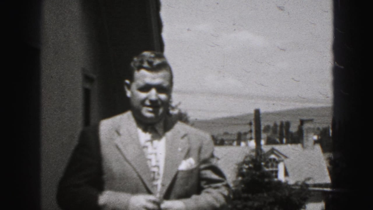 Suited man facing the camera outdoors with rooftops and distant hills behind him.