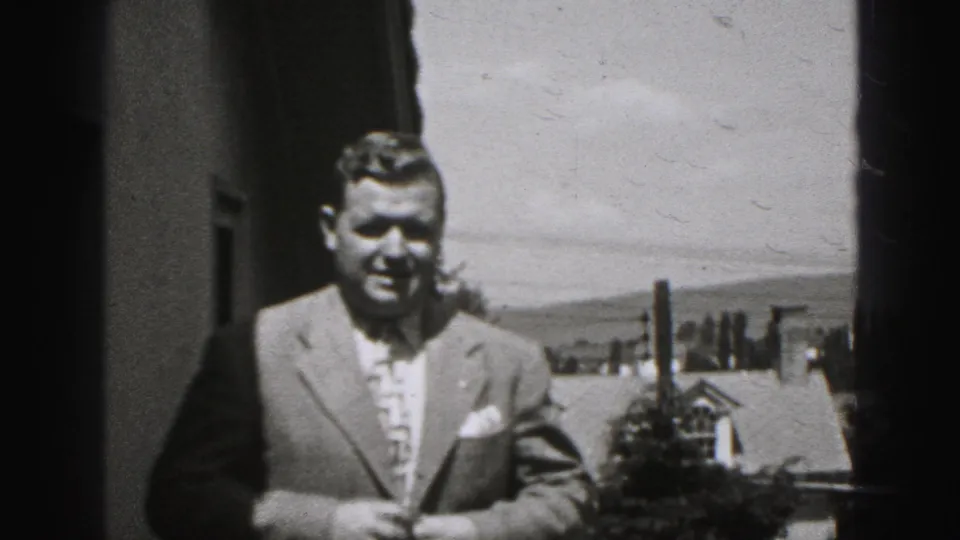 In metadata-supported 1975 Harrisburg footage, a suited adult man stands outdoors facing the camera with rooftops, chimneys, trees, and hills behind him.