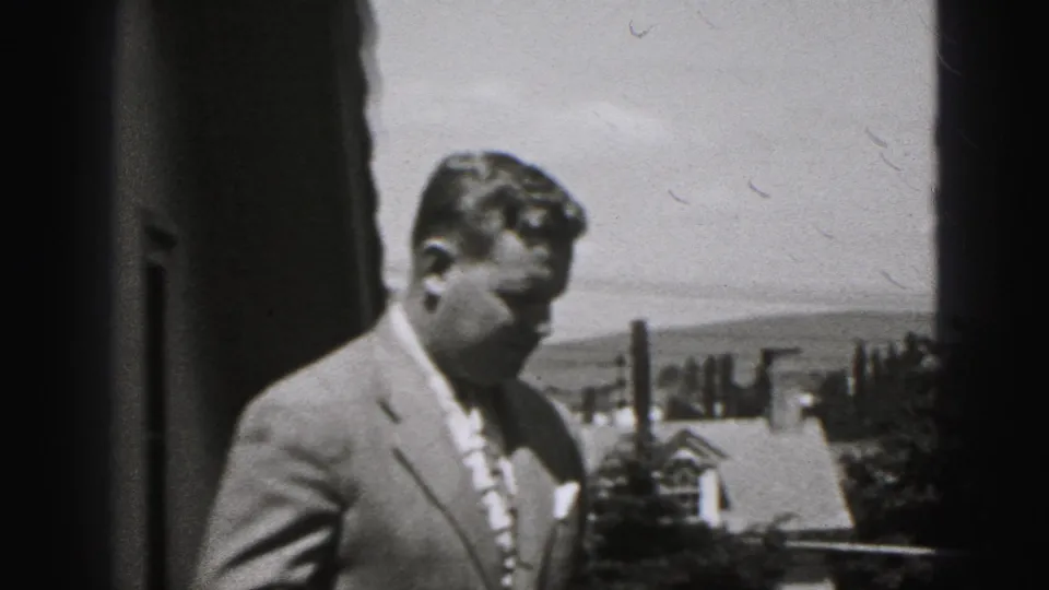 The man turns sideways in the 1975 Harrisburg home-movie frame, with a suburban-looking roofline, chimney forms, trees, and hills visible behind him.