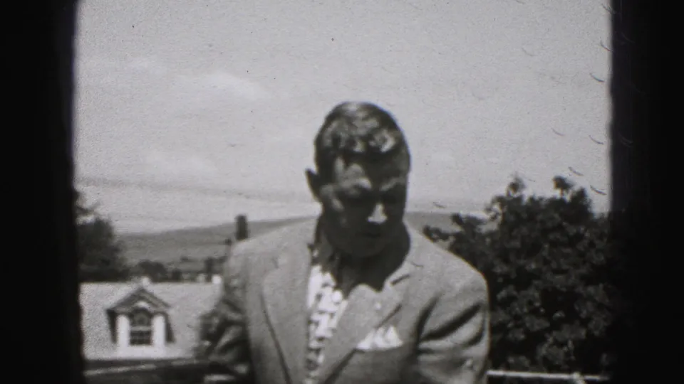 A suited adult man looks down in the Harrisburg-area 1975 frame, partly shadowed, with a roof dormer, trees, and open sky behind him.