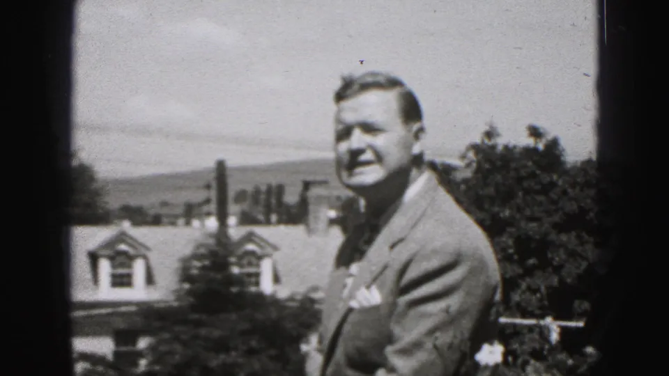 In the final 1975 Harrisburg frame, the suited man turns toward the camera near rooftops, trees, chimneys, distant hills, and bright sky.
