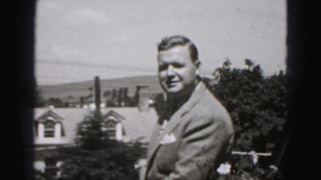 Adult man in a suit smiles outdoors near shrubs, a house roofline, and distant hills.