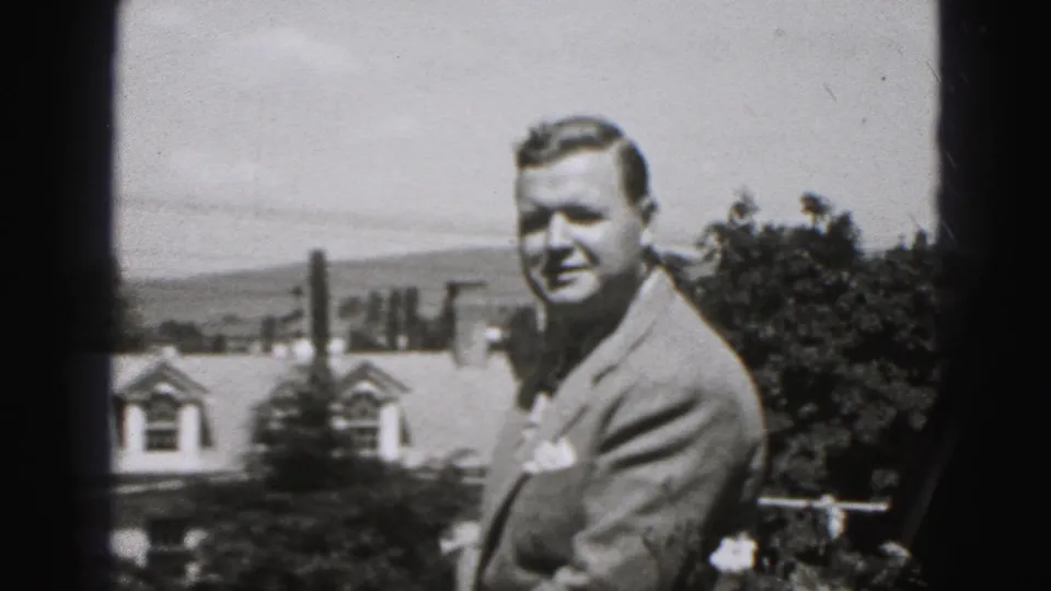 In 1975 Harrisburg home-movie footage, an adult man in a light suit smiles toward the camera in a garden or yard setting.