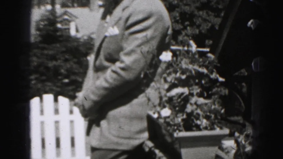 The frame shows the man's torso in a suit beside a white picket fence, shrubs, and flowers in the outdoor Harrisburg setting.